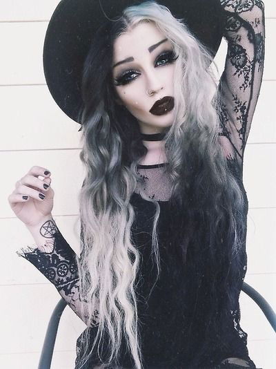 A trendy Goth look with dark lips, black and white long hair, lace top and a big, black hat.